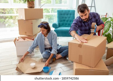 Couple In Love Packing Things Into Cardboard Boxes, Getting Ready For Relocation - Man Taping Boxes Using Packing Machine While Woman Is Wrapping Fragile Things Into Paper