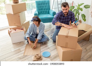 Couple In Love Packing Things Into Cardboard Boxes, Getting Ready For Relocation - Man Taping Boxes Using Packing Machine While Woman Is Wrapping Fragile Things Into Paper