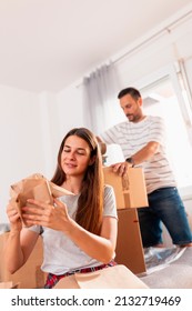 Couple In Love Packing Things Into Cardboard Boxes, Getting Ready For Relocation - Man Taping Boxes Using Packing Machine While Woman Is Wrapping Fragile Things Into Paper