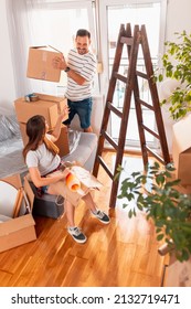 Couple In Love Packing Things Into Cardboard Boxes, Getting Ready For Relocation - Man Taping Boxes Using Packing Machine While Woman Is Wrapping Fragile Things Into Paper