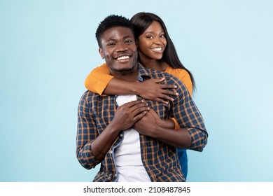Couple In Love. Portrait of joyful black woman hugging her boyfriend from behind, standing together isolated over blue studio background. Casual guy and lady smiling, posing and looking at camera - Shutterstock ID 2108487845