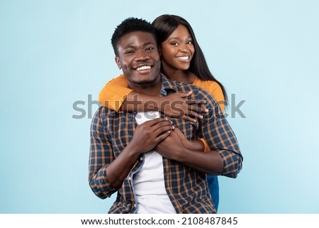 Couple In Love. Portrait of joyful black woman hugging her boyfriend from behind, standing together isolated over blue studio background. Casual guy and lady smiling, posing and looking at camera