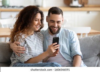 Couple in love resting on comfy couch having fun using smart phone apps, enjoy distant video call, wife showing interesting website to husband, choosing services goods on-line e-commerce usage concept