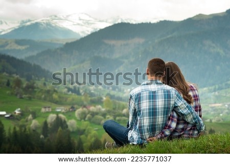 A couple in love sit in embraces on the top of mountain with a scenic view on the background. 
