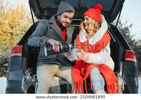 Couple in love sitting in car trunk drinking hot tea in snowy winter forest and chatting. People relaxing outdoors during road trip. Valentines day
