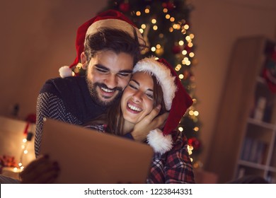 Couple in love sitting next to a Christmas tree, wearing Santa hats and having fun looking at old photos on a tablet computer