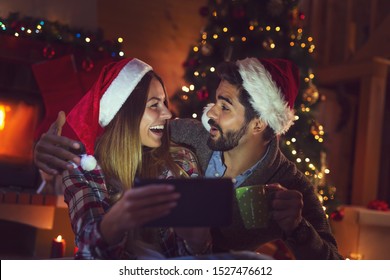 Couple in love sitting next to a Christmas tree and a fireplace, wearing Santa hats and having fun looking at old photos on a tablet computer - Powered by Shutterstock