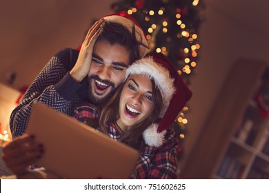 Couple in love sitting next to a Christmas tree, wearing Santa's hats and having fun looking at old photos on a tablet computer. Focus on the girl