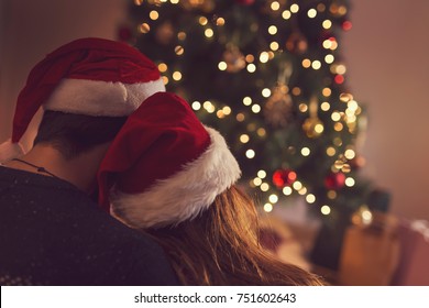 Couple in love sitting next to a Christmas tree, wearing Santa's hats, hugging and looking away from the camera towards the tree. Selective focus on the girl's hat - Powered by Shutterstock