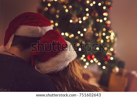 Couple in love sitting next to a Christmas tree, wearing Santa's hats, hugging and looking away from the camera towards the tree. Selective focus on the girl's hat