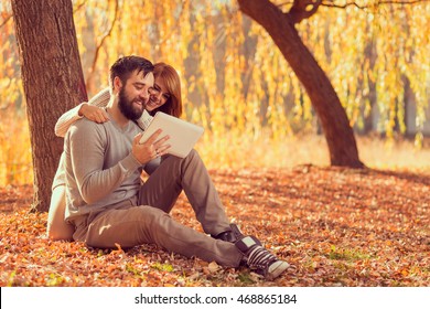 Couple in love sitting on autumn fallen leaves in a park, surfing the net on a tablet computer in search for travel destinations. Focus on a guy - Powered by Shutterstock