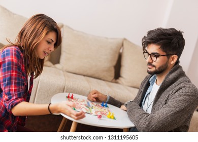 Couple In Love Sitting On The Floor Next To A Table, Playing Ludo Board Game And Enjoying Their Free Time Together. Woman Rolling The Dice