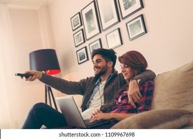 Couple in love sitting on a living room sofa, watching TV. Man using a remote control while woman is surfing the net on a laptop computer. Focus on the man