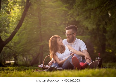 Couple In Love Sitting On A Picnic Blanket In A Park, Talking To Each Other, Drinking Wine And Enjoying A Beautiful, Peaceful Day In Nature