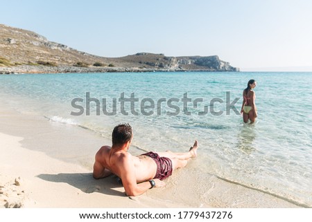 Couple in love spends leisure time together on the beach. A guy lies on the sand and looks how his girlfriend swims