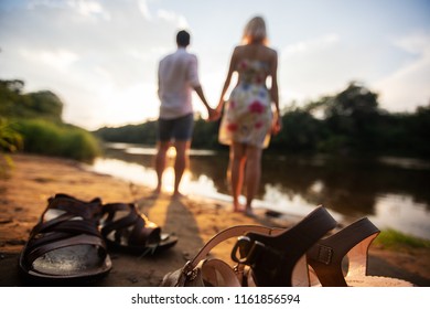 A Couple In Love Stands On The Shore Of A Pond, A River, A Lake, The Sea Or The Ocean, Trying To Kick The Feet With Water, They Take A Bath, Take Off Their Shoes, Put It On A Sandy Beach, Summer Sunny