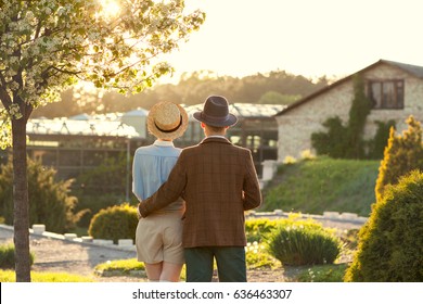 A Couple In Love At Sunset In Hats Stand With Their Backs In The Frame. American Dream. A Retro Couple In His Country House, His Estate. Lavstory, Family Day, St. Valentine's Day.