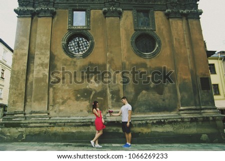 Couple in love are walking in the old ancient town.  Man in shorts is giving flowers to his girl in pink dress near the medieval stone church. Romantic summer hipster stylish photo. Shy emotions.