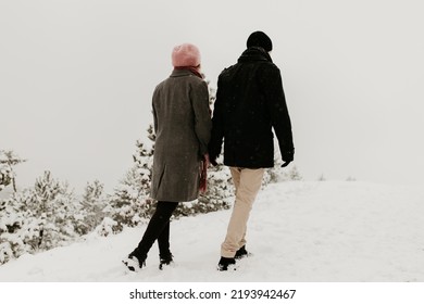 A Couple In Love Is Walking In The Snow In The Mountains. Winter Outdoor Hiking Sport Activity. Warm Coat, Knitted Cap And Boots On Them. Holding Each Others Hand. Romantic Walk In The Hills. 