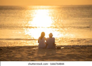 Couple In Love Watching Sunset Together On Beach Travel Summer Holidays. People Silhouette From Behind Sitting Enjoying View Sunset Sea On Tropical Destination Vacation. Romantic Couple On The Beach.