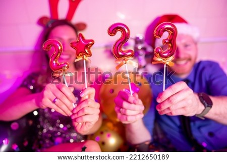 Couple in love wearing Santa hat and antlers sitting in a bathtub filled with balloons and confetti having fun at New Year Eve party, holding balloons shaped as numbers 2023