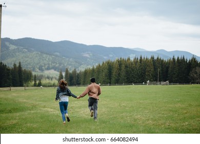 Couple Of Lovers Holding Hands And Running Away In Mountains. Woman And Man In Love Walking Outdoors. Couple In Checkered Shirts.