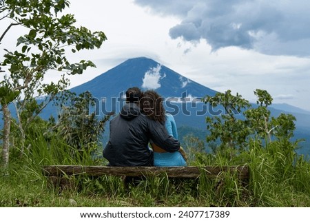 A couple of lovers are sitting on a bench, hugging each other and enjoying the view of the popular sacred cloud-covered Mount Agung on the island of Bali