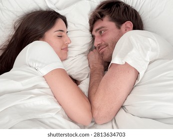 Couple lying in bed facing each other, smiling and cuddling under white sheets, intimate morning moment conveying love, comfort, relaxation and connection