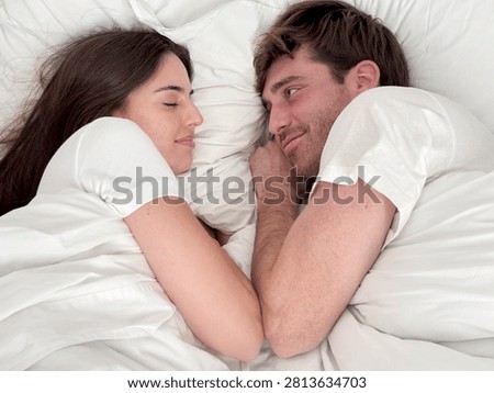 Couple lying in bed facing each other, smiling and cuddling under white sheets, intimate morning moment conveying love, comfort, relaxation and connection