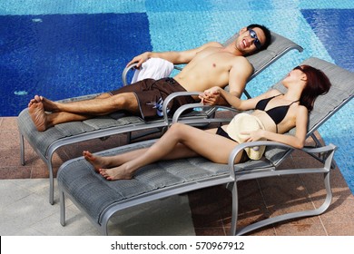 Couple Lying On Deck Chairs By Pool, Holding Hands