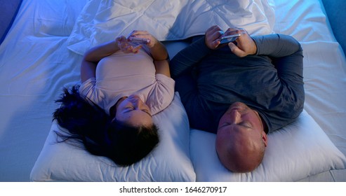 A Couple Is Lying On White Bed Linen With Smartphones In Their Hands. A Woman With Long Hair Asks A Question, A Bald Man Shows Her Something On His Phone. The View From The Top. Blue Bottom Light