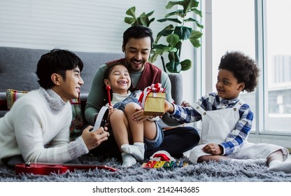 Couple male gay taking care, giving gift to adopted children who are little diverse Caucasian and African girl and boy with happiness to celeblate birthday in living room at home. LGBT, kids Concept