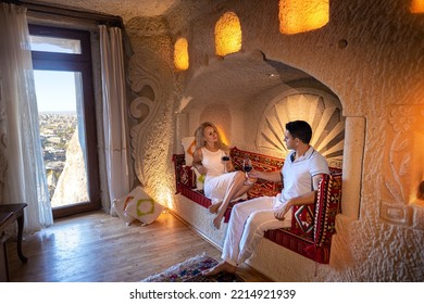 A Couple Man And Woman Drinking Red Wine In The Old Cave Room Of Cappadocia On The Carved Bed With Beautiful Window View