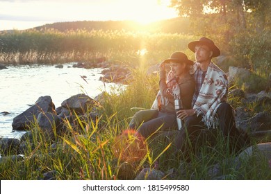 Couple Man And Woman In Felt Hats Wrapped In Whool Blanket Sitting Near Lake On Sunset. Weekend Picnic On Nature, Local Travel, Staycation, Folk Lifestyle. 