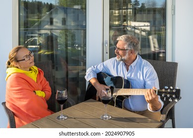 Couple man and woman relaxing outside the house on terrace or balcony. Mid aged female and senior male smiling enjoying red wine in glasses. Bearded man imposing musician plays the guitar and sings. - Powered by Shutterstock