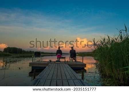 Couple man and woman seated on a wooden jetty, looking a colorful sunset on the lake and fishing