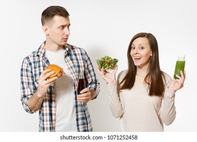 Couple Man And Woman Standing With Green Detox Smoothies, Salad In Glass Bowl, Burger, Cola In Glass Bottle Isolated On White Background. Proper Nutrition, Healthy Lifestyle, Fast Food Choice Concept