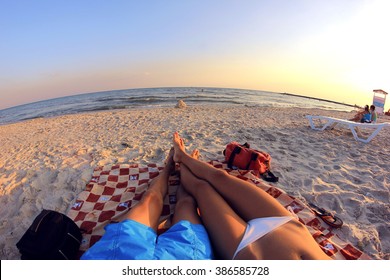 Couple (man And Woman) Sunbathing On The Beach Overlooking The Sea. They Lie On A Mat On The Sand. In Front Of Them A House Of Sand And Water. Around Other People Are Resting. Ukraine, Black Sea Coast