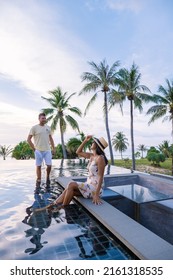 Couple Man And Woman Watching Sunset From A Rooftop Infinity Pool On A Luxury Vacation. European Men And Asian Woman Mid Age Watch Sunset At The Pool 