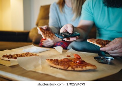 A Couple Of Men And Women Eat Pizza On The Sofa In Their Living Room At Night While Watching A Movie On Television. Concept Of Life As A Couple At Home.