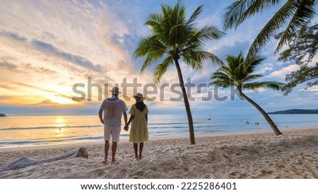 A couple of men and women standing by a palm tree watching the sunset on the beach with white sand and palm trees, Bang Tao beach Phuket Thailand. 