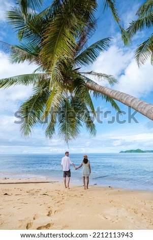a couple of men and women walking under a hanging palm tree on vacation at a tropical Island in Thailand Koh Mak. turquoise colored water and palm trees at a tropical island, drone aerial view