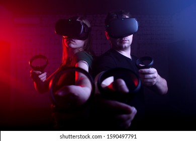 Couple In Modern Wireless Glasses Of Virtual Reality Play A Shooter Against A Dark Neon Background, A Team Of Gamers In A Game With Weapons