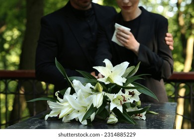 Couple near granite tombstone with white lilies at cemetery outdoors, selective focus. Funeral ceremony