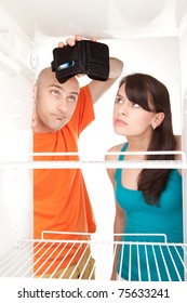 A Couple With No Money Searching For Food In An Empty Refrigerator Whilst The Man Holds His Empty Wallet.