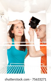 A Couple With No Money Searching For Food In An Empty Refrigerator Whilst The Man Holds His Empty Wallet.