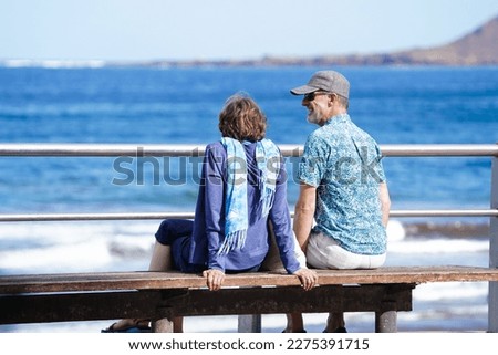 Couple of old retired seniors man and woman siting at a bench watching ocean by the beach and laughind or smiling