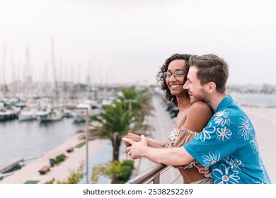 A couple is on a balcony overlooking a marina. The man is wearing a blue shirt with flowers. The woman smiles and holds the man's hand. The scene is romantic and peaceful. - Powered by Shutterstock