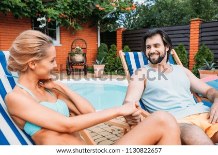 couple on beach chairs holding hands while spending time near swimming pool on backyard on summer day