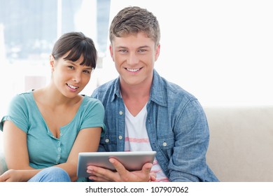 A Couple On The Couch With A Tablet Pc Smiling As They Look Straight Ahead Into The Camera